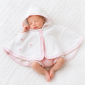 A sleeping newborn baby wearing a white hooded blanket cape with pink trim and matching pink footed diaper cover, lying on a white textured blanket.