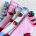 Four pairs of knit baby socks arranged diagonally, featuring strawberry patterns in pink, white, and blue. The pink pair has red and green strawberries, the white pair has red and green strawberries, and the blue-and-white striped pair has blue and green strawberries.