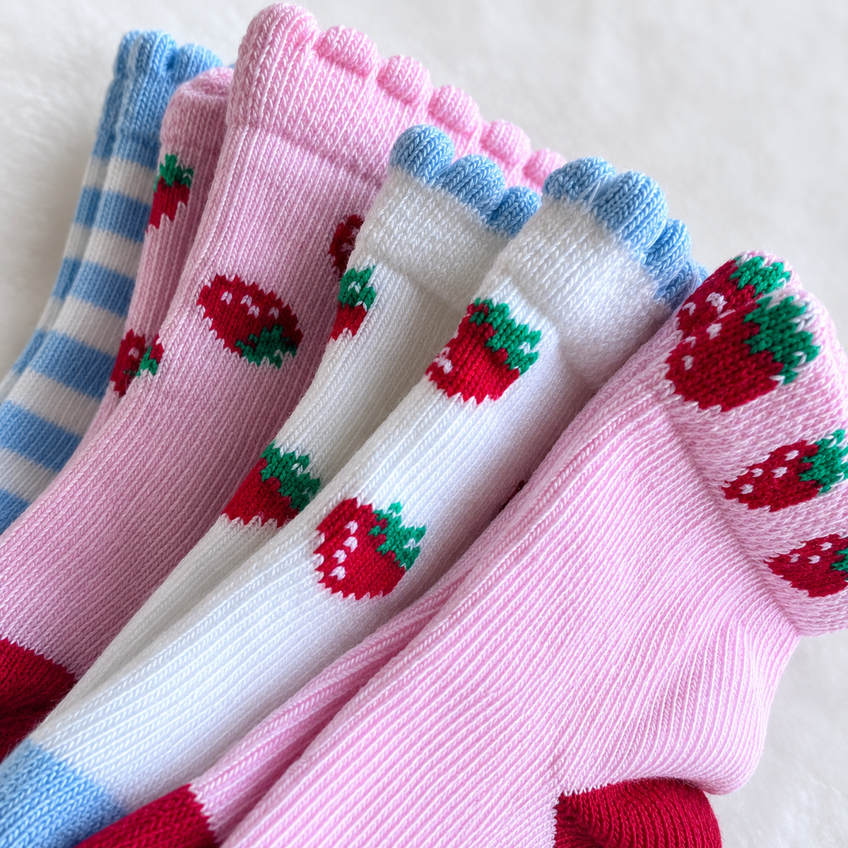 Four pairs of knit baby socks arranged diagonally, featuring strawberry patterns in pink, white, and blue. The pink pair has red and green strawberries, the white pair has red and green strawberries, and the blue-and-white striped pair has blue and green strawberries.