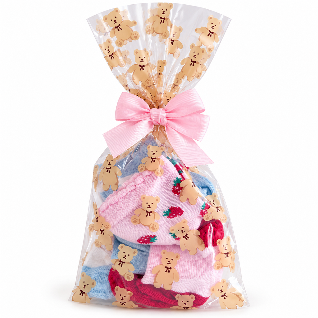A clear cellophane gift bag filled with folded pink and blue knit socks, tied with a pink ribbon at the top, and decorated with repeated teddy bear cutouts on a white background.
